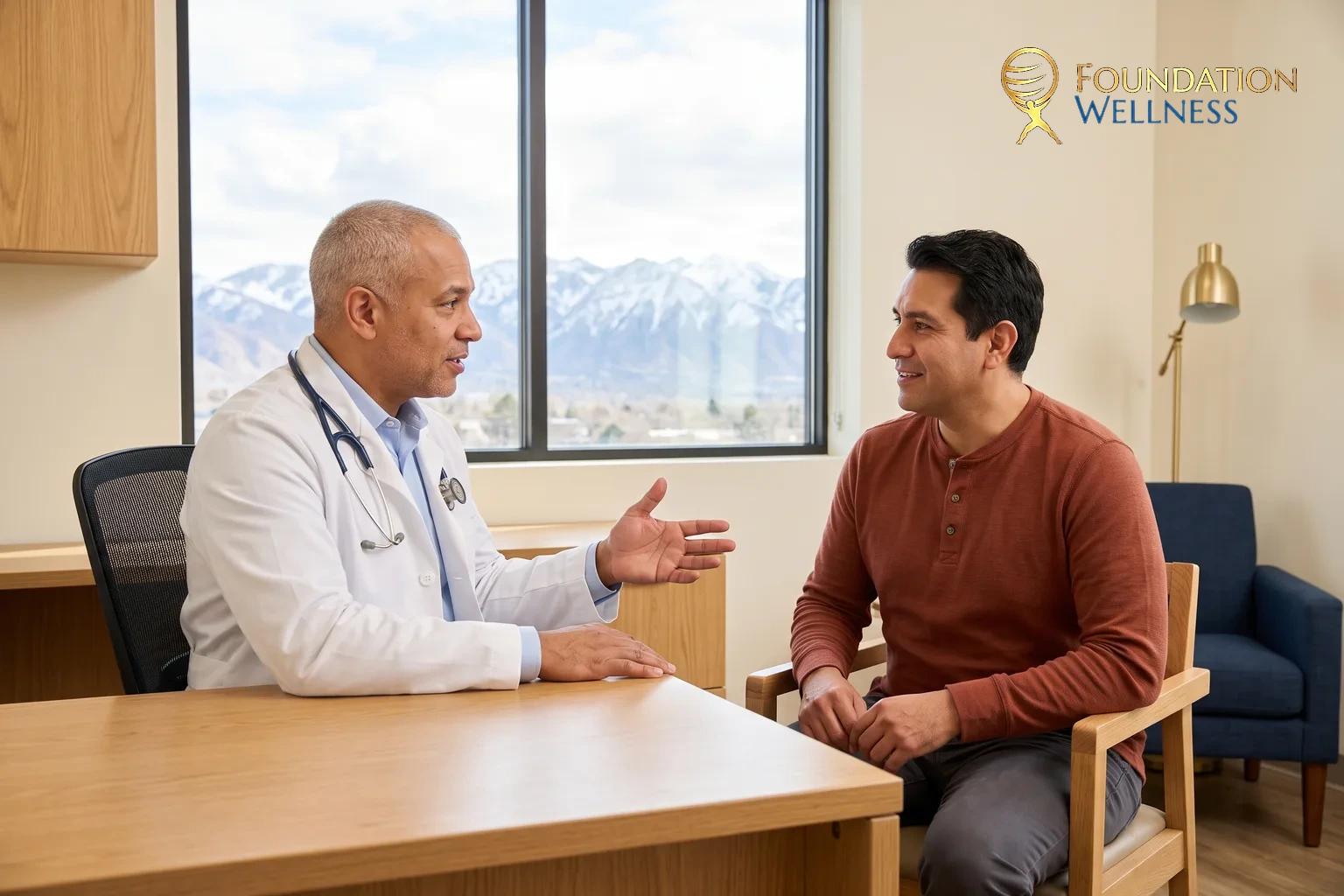 A physician in a white coat with a stethoscope talking across a light oak desk with a man in a rust henley, snow-topped mountains through the window